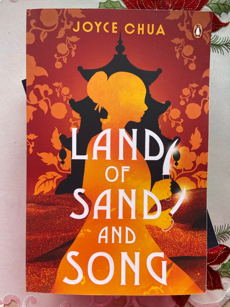 Land of Sand and Song by Joyce Chua is on a white cloth with embroidered red flowers